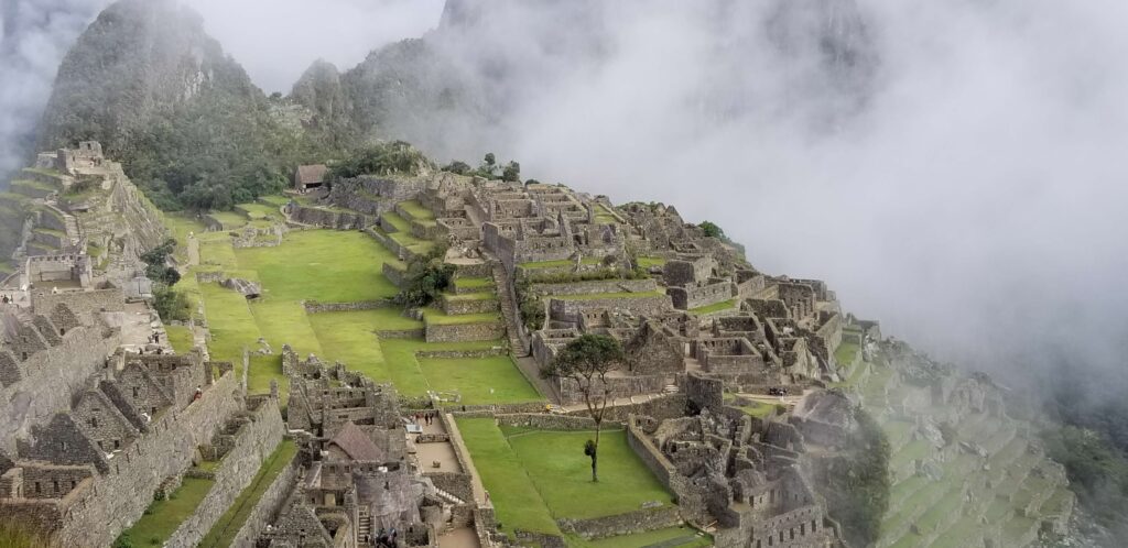 Wide-angle landscape view of the ancient Incan citadel of Machu Picchu in Peru, featuring stone ruins, terraced fields, and lush green mountains partially shrouded in low-lying white clouds.